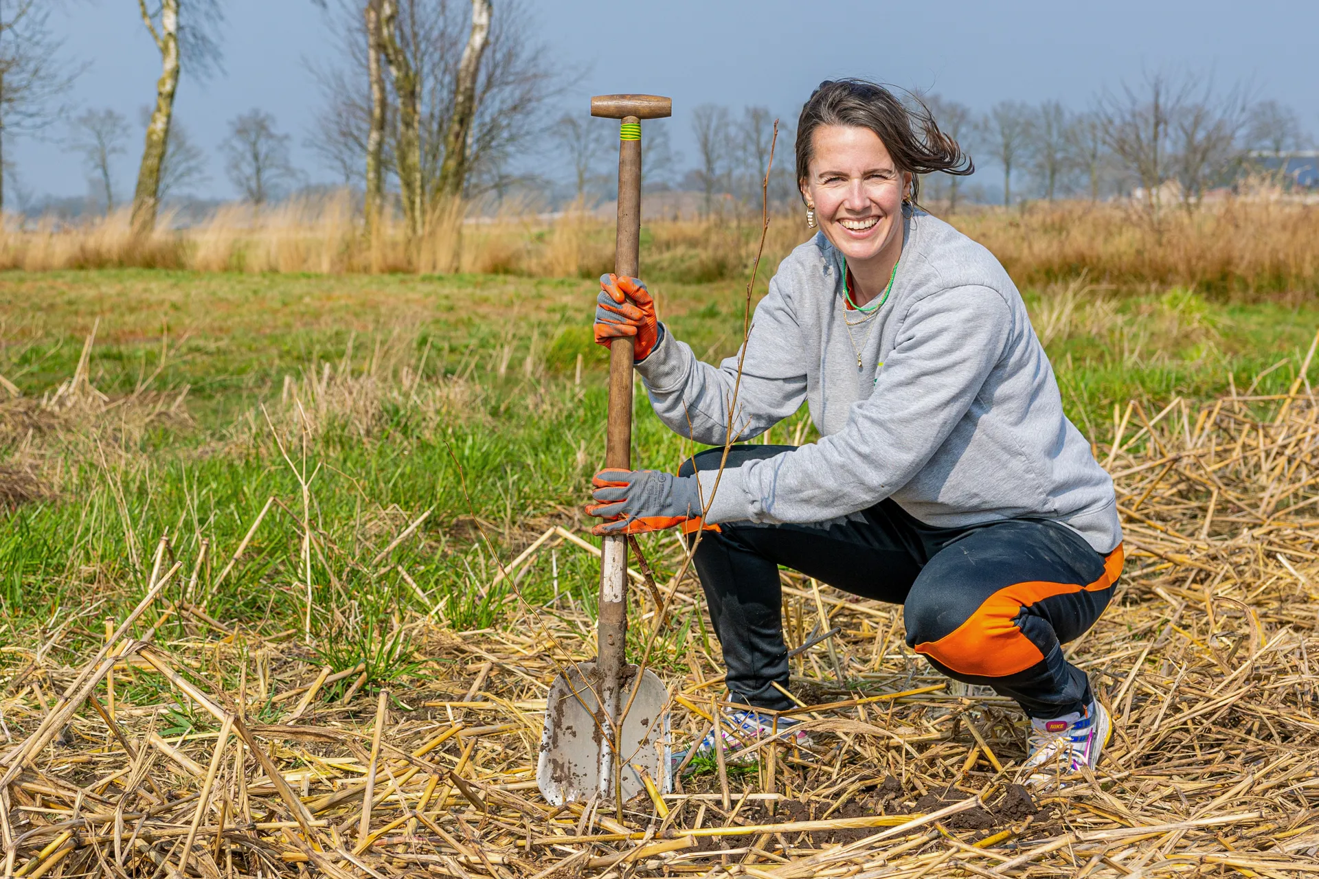 Margriet Klapwijk Een vrouw in casual tuinkleding plant een jonge boom op een open grasveld. Ze hurkt naast een boom in de grond en houdt een schep vast.