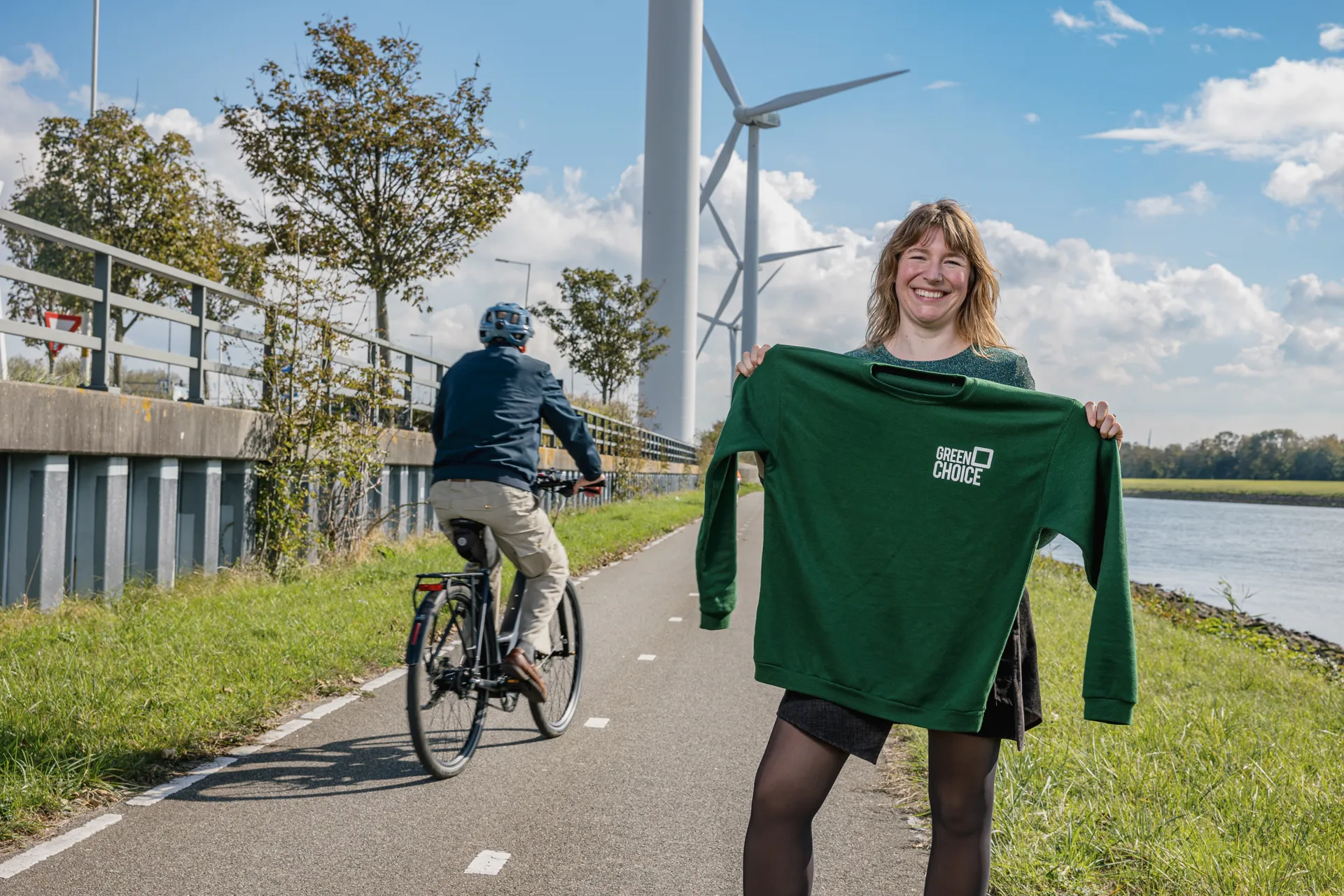 Vrouwelijke collega staat lachend met een Greenchoice trui in haar hand. Ze is buiten en staat langs een fietspad met windmolens op de achtergrond. Er rijdt een fietser langs haar