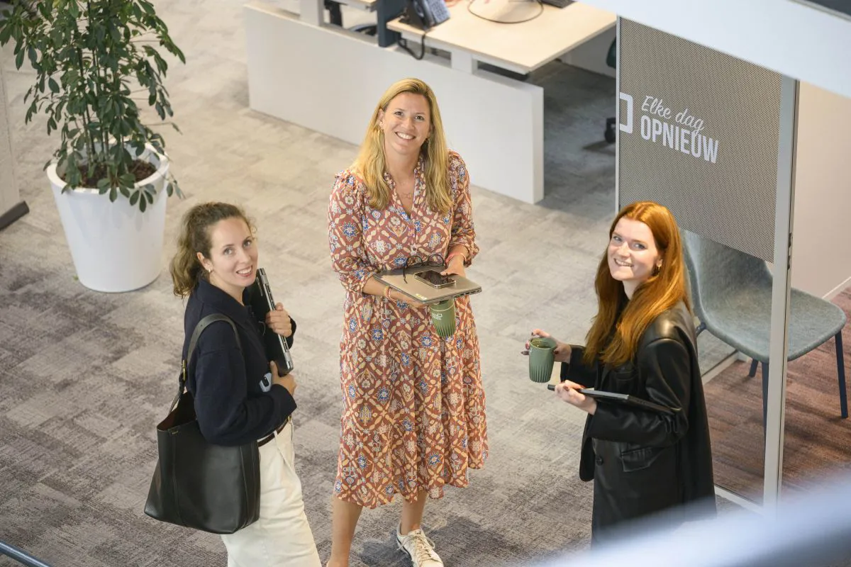 3 vrouwelijke collega's kijken in de camera. Zij kijken al lachend in de camera en staan te wachten voor een meetingruimte.