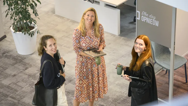 3 vrouwelijke collega's kijken in de camera. Zij kijken al lachend in de camera en staan te wachten voor een meetingruimte.