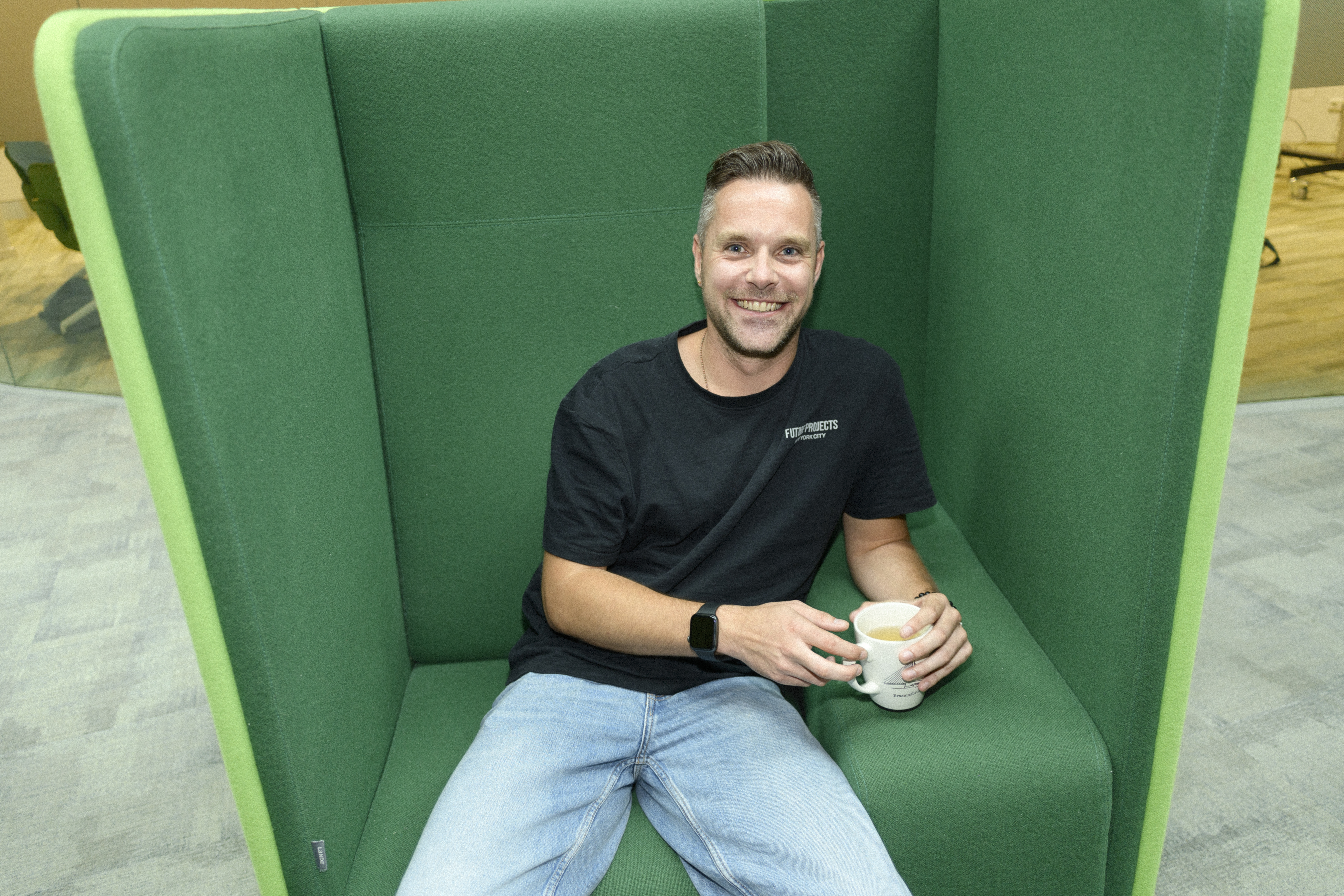 Een man zit ontspannen in een comfortabele stoel, met een rustige en tevreden uitstraling. Hij leunt iets achterover met een kop koffie in zijn handen.