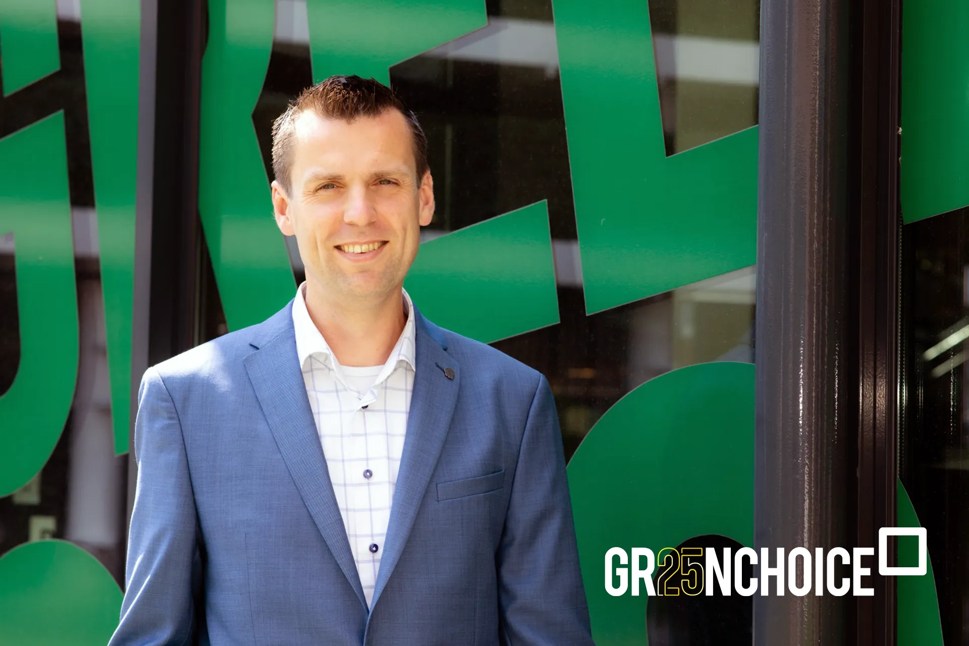 GC 25 Maurice Man (Maurice) kijkt lachend in de camera. De zon schijnt op zijn gezicht terwijl hij buiten voor een raam met het Greenchoice logo staat.