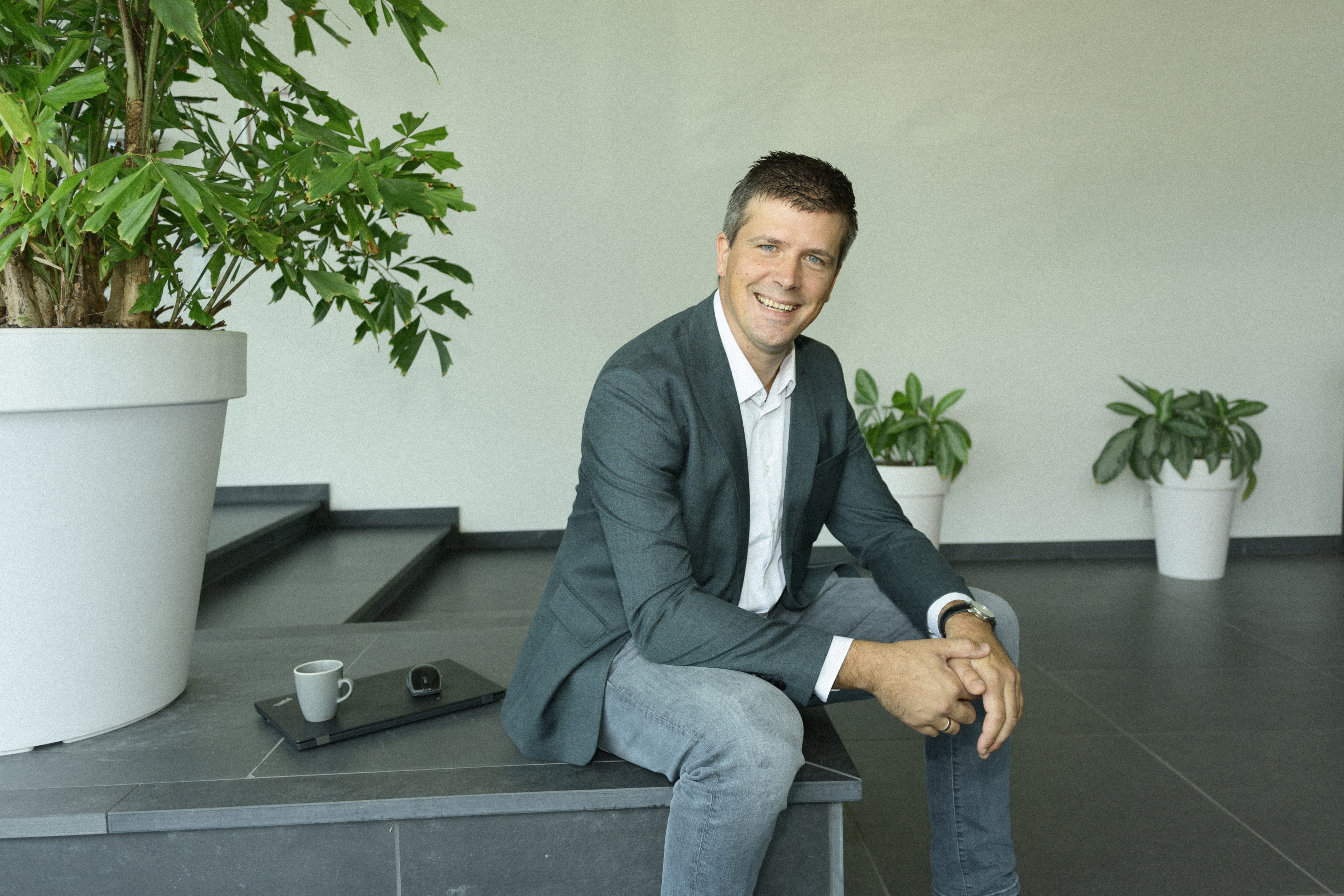 Een man zit ontspannen op een stenen verhoging met achter zich een dichtgeklapte laptop. Hij kijkt recht in de camera en lacht vriendelijk. 