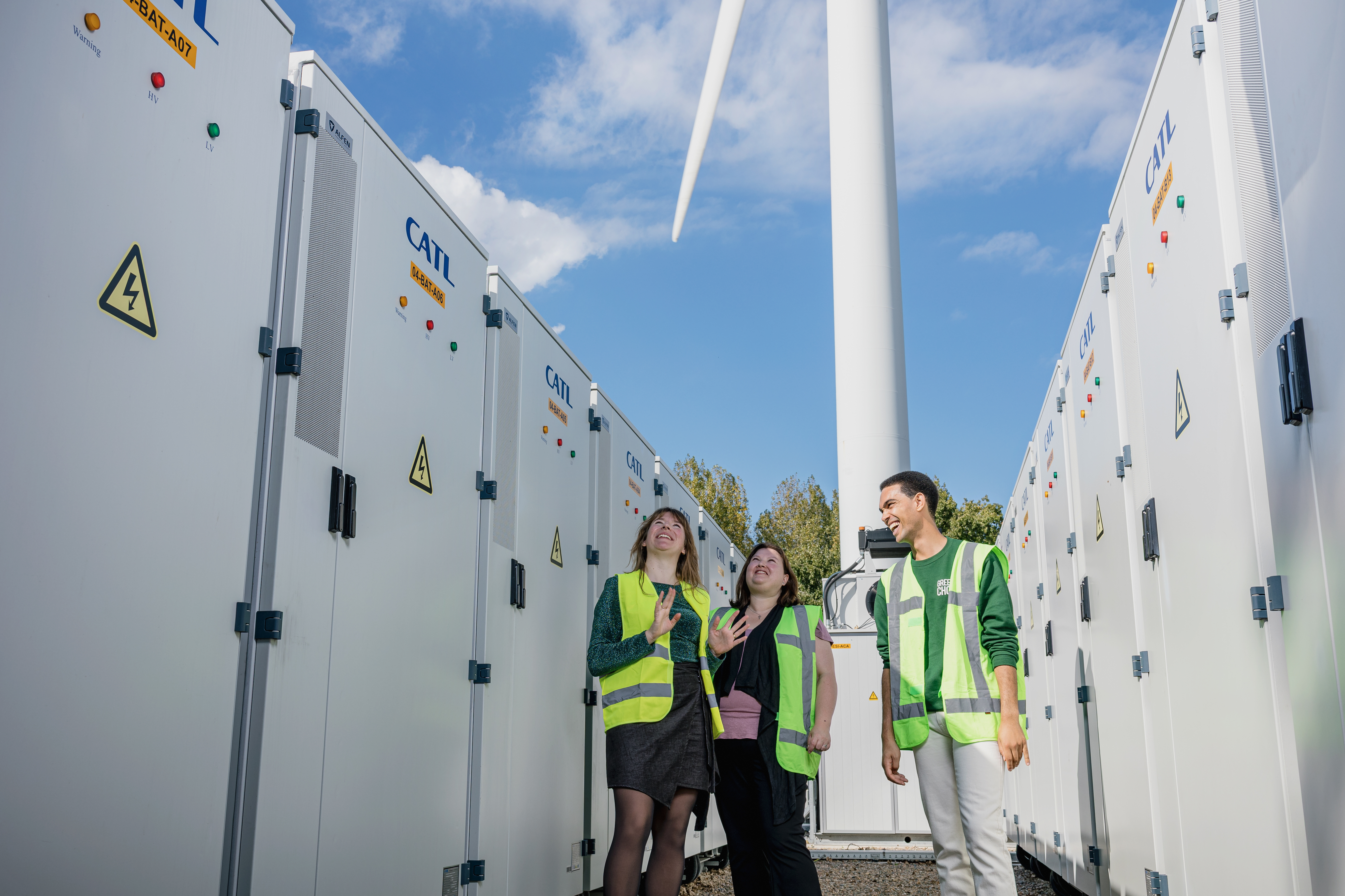 Drie collega's staan lachend en omhoog kijkend tussen grote batterijen met een windmolen op de achtergrond onder een heldere blauwe lucht