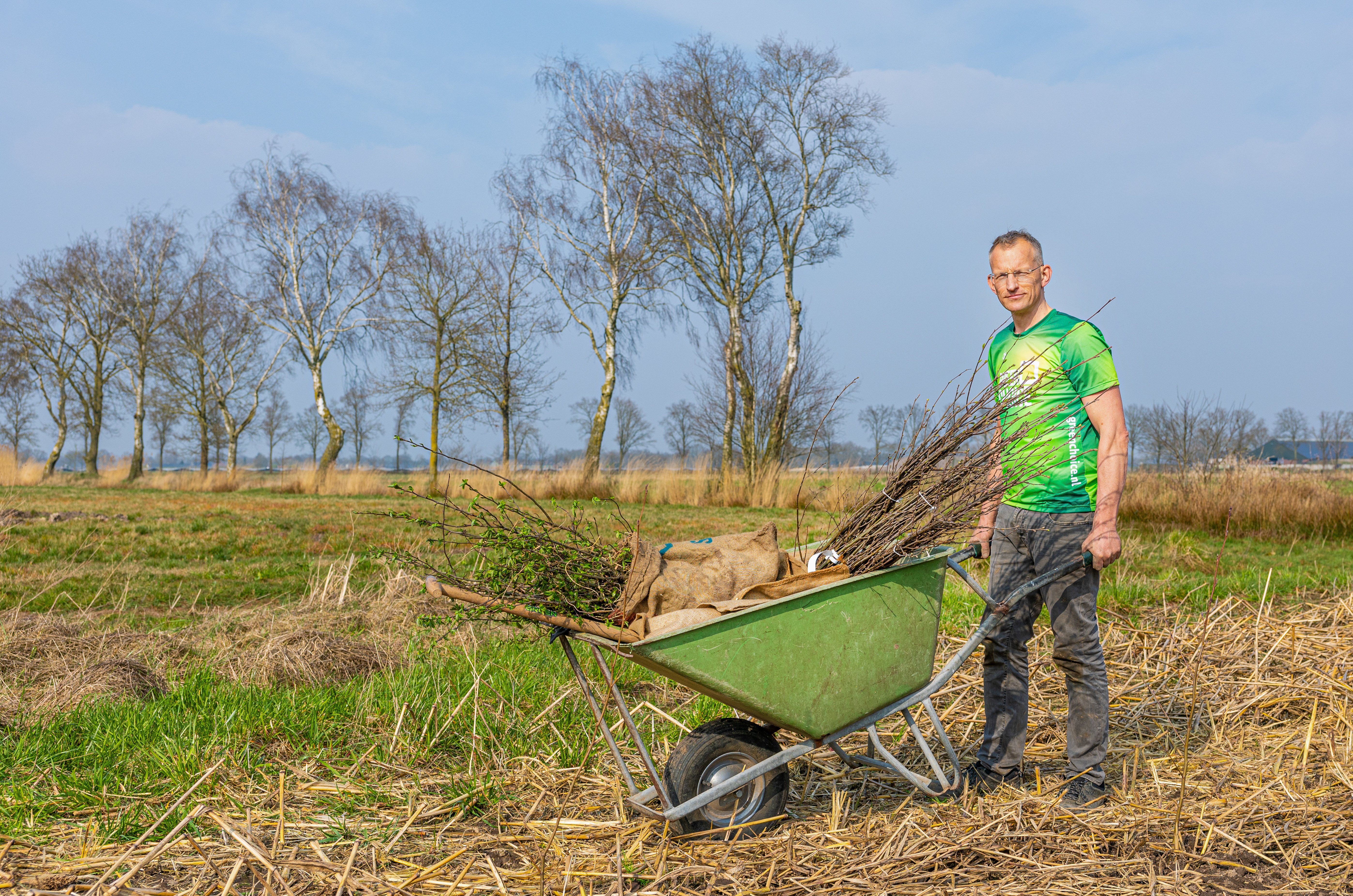 Een man houdt een kruiwagen vol jonge boompjes vast in een groene, natuurlijke omgeving. Hij staat op een grasveld klaar om bomen te planten. Op de achtergrond zijn hoge bomen en struiken zichtbaar.