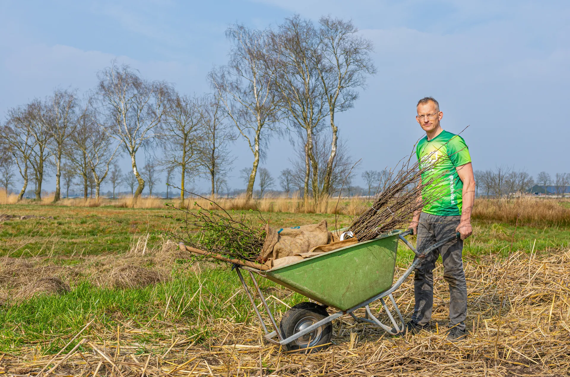 Jacobus van der Hek Een man houdt een kruiwagen vol jonge boompjes vast in een groene, natuurlijke omgeving. Hij staat op een grasveld klaar om bomen te planten. Op de achtergrond zijn hoge bomen en struiken zichtbaar.