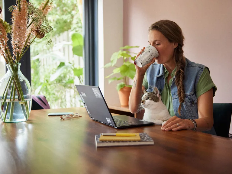 Vrouw met kat aan computer Een vrouw drinkt uit een kop terwijl ze naar haar laptop kijkt. Ze zit aan een eettafel. Een kat zit op schoot en kijkt ook naar de laptop.