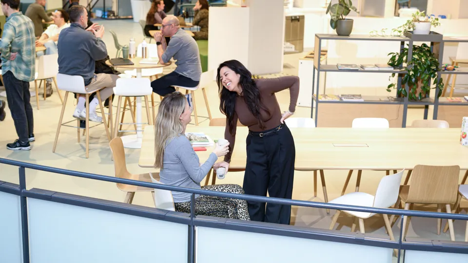 Twee vrouwen zitten ontspannen en lachend in een open kantoorruimte, omringd door natuurlijk licht en een moderne inrichting. Hun houding straalt collegialiteit en plezier uit. Een van de vrouwen is zwanger.