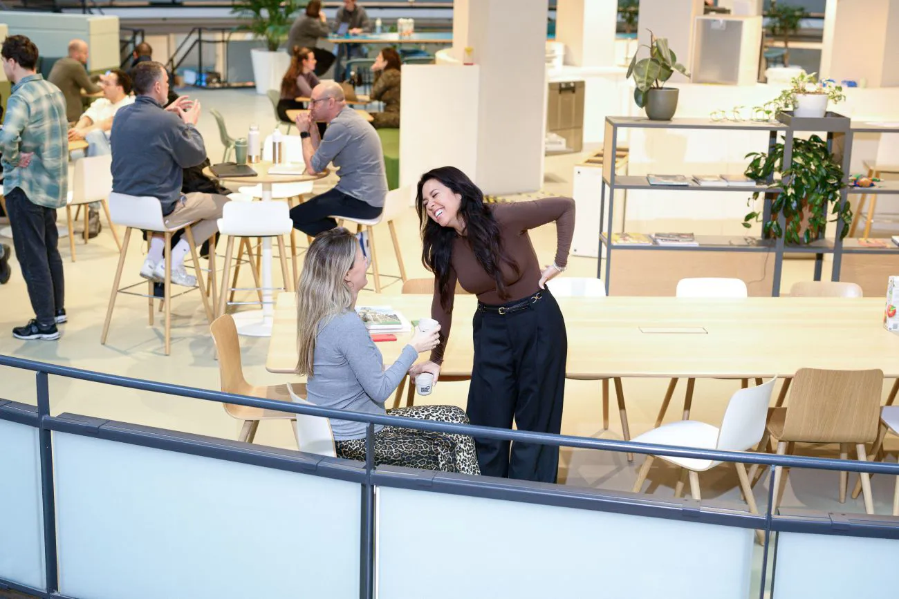 Twee vrouwen zitten ontspannen en lachend in een open kantoorruimte, omringd door natuurlijk licht en een moderne inrichting. Hun houding straalt collegialiteit en plezier uit. Een van de vrouwen is zwanger.