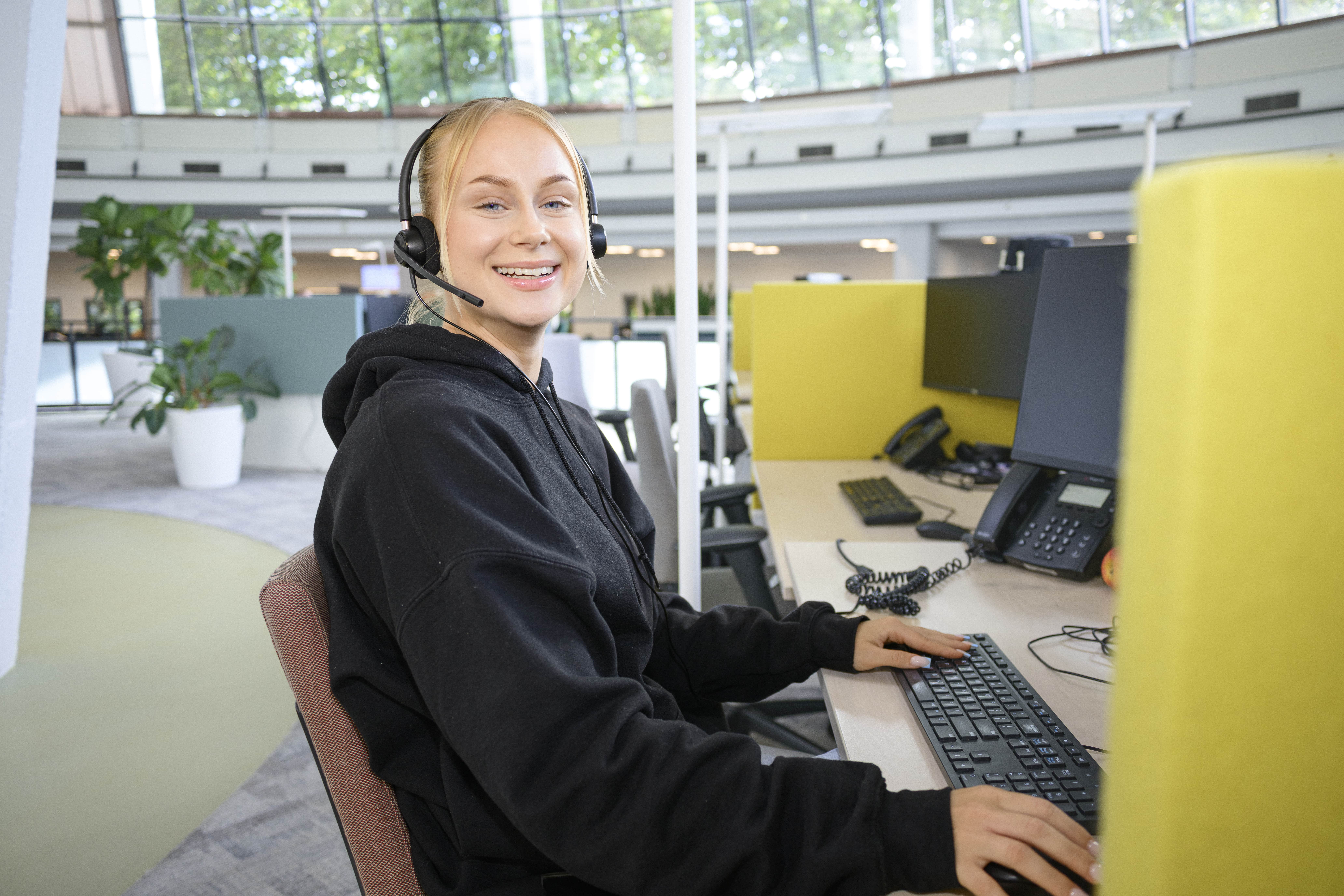 Collega zit met headset op aan haar werkplek. Ze kijkt lachend in de camera en geeft een ontspannen indruk in haar hoodie. 