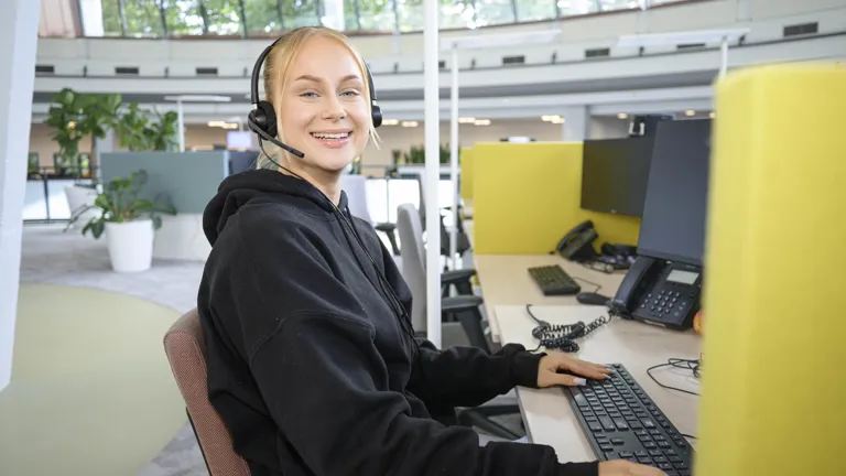 Collega zit met headset op aan haar werkplek. Ze kijkt lachend in de camera en geeft een ontspannen indruk in haar hoodie.