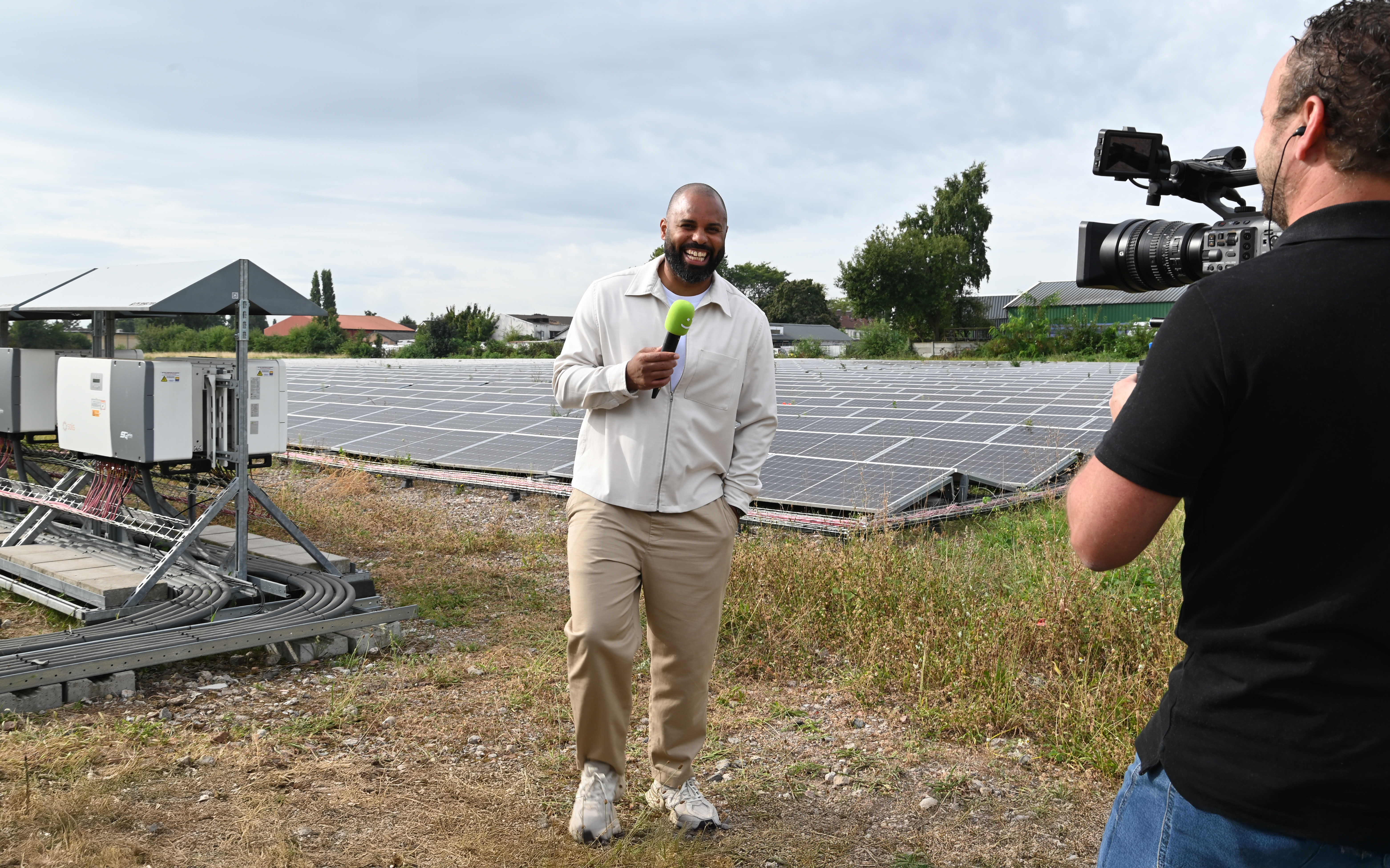 Edi staat op een veld vol zonnepanelen. Hij wordt gefilmd en houdt lachend een microfoon in in zijn hand. Het is mooi weer en hij staat er ontspannen bij. 