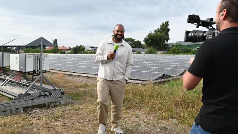 Edi staat op een veld vol zonnepanelen. Hij wordt gefilmd en houdt lachend een microfoon in in zijn hand. Het is mooi weer en hij staat er ontspannen bij.