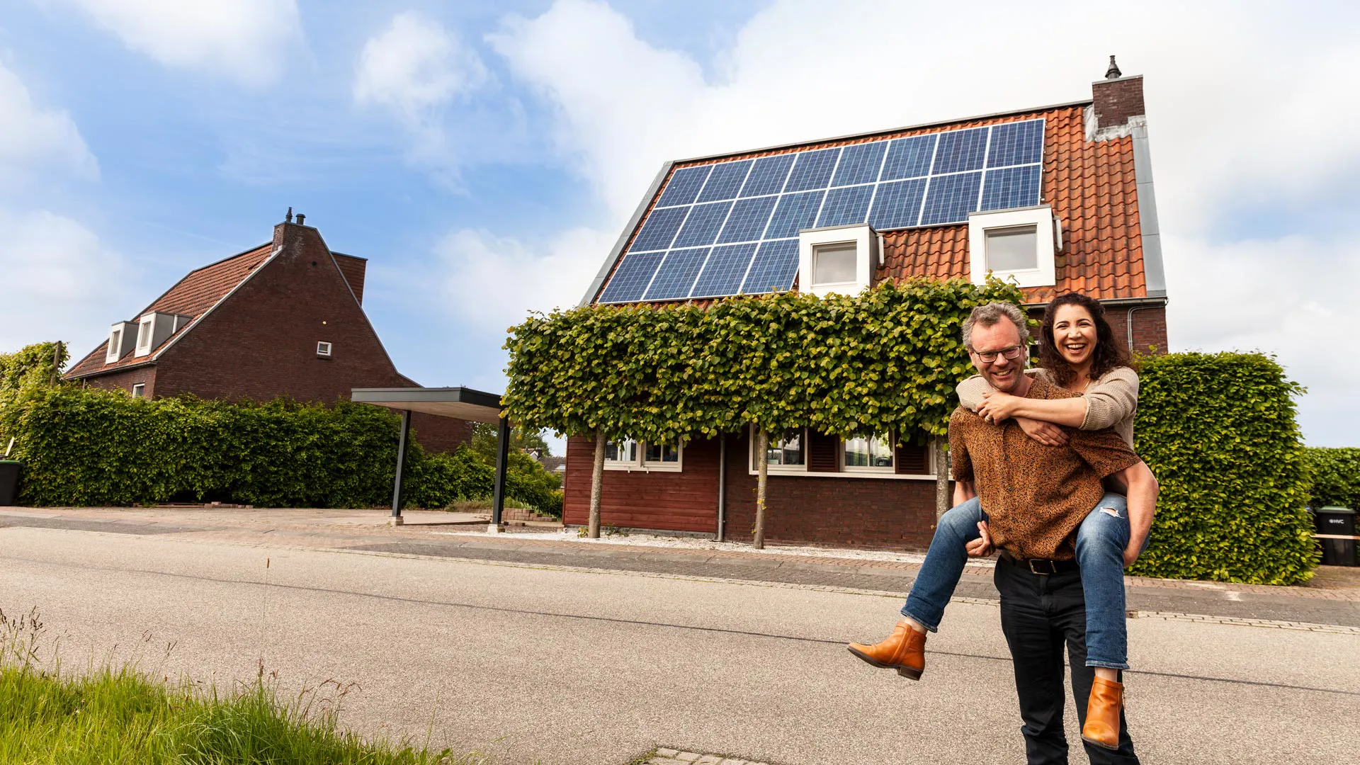 Koppel voor huis met zonnepanelen Iemand zit op de rug van een man. Samen kijken ze in de camera. In de achtergrond staat een woning met zonnepanelen die geplaatst zijn door Greenchoice.