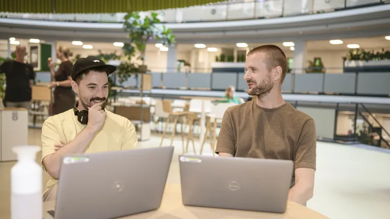 Twee collega's hebben een informeel overleg met de laptop in het Groene Hart (onze lunchruimte). Ze ogen ontspannen en kijken naar elkaar of het scherm.