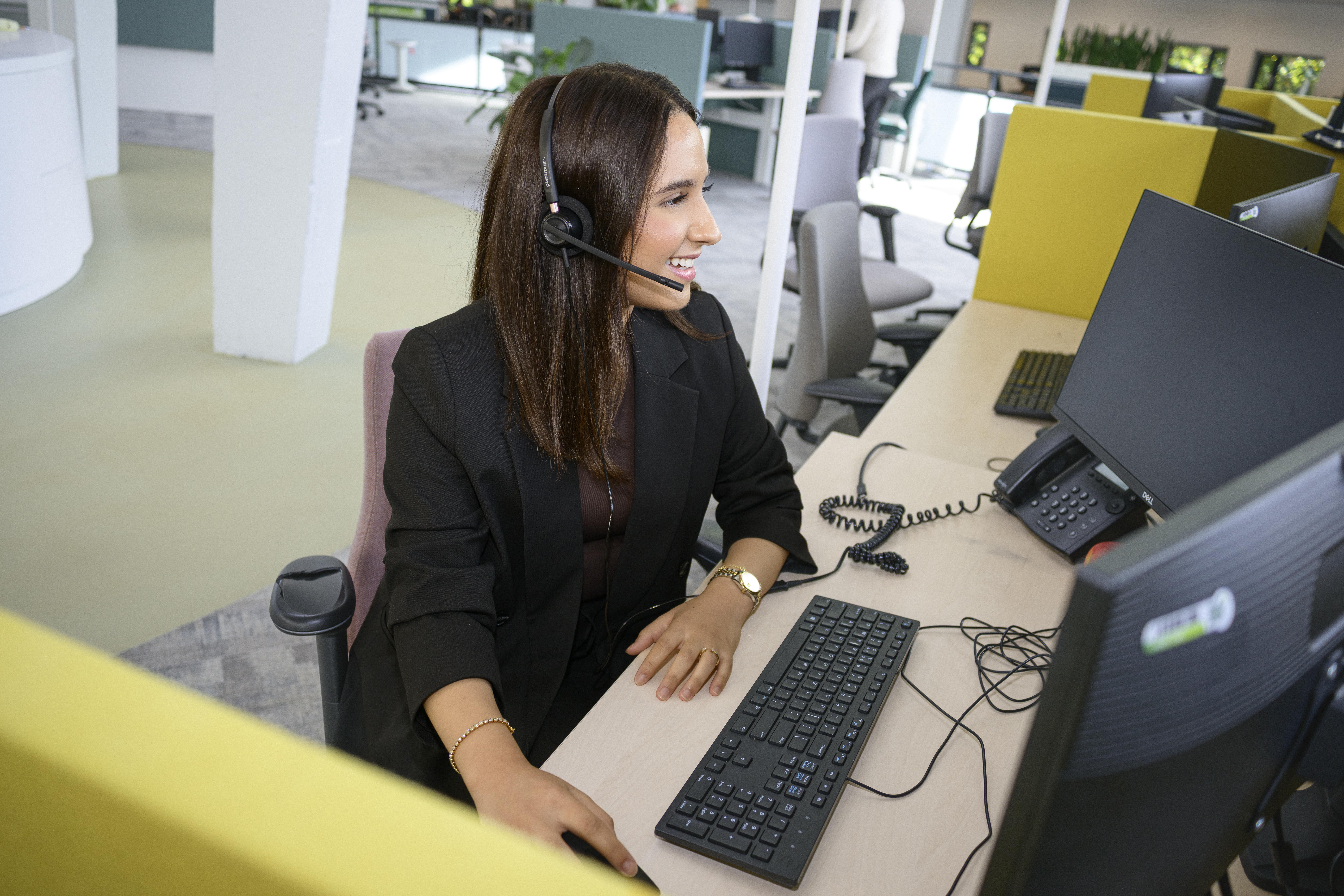 Collega zit op haar werkplek. Ze is aan het werk met een headset op haar telefoon waarmee ze klanten te woord staat. 