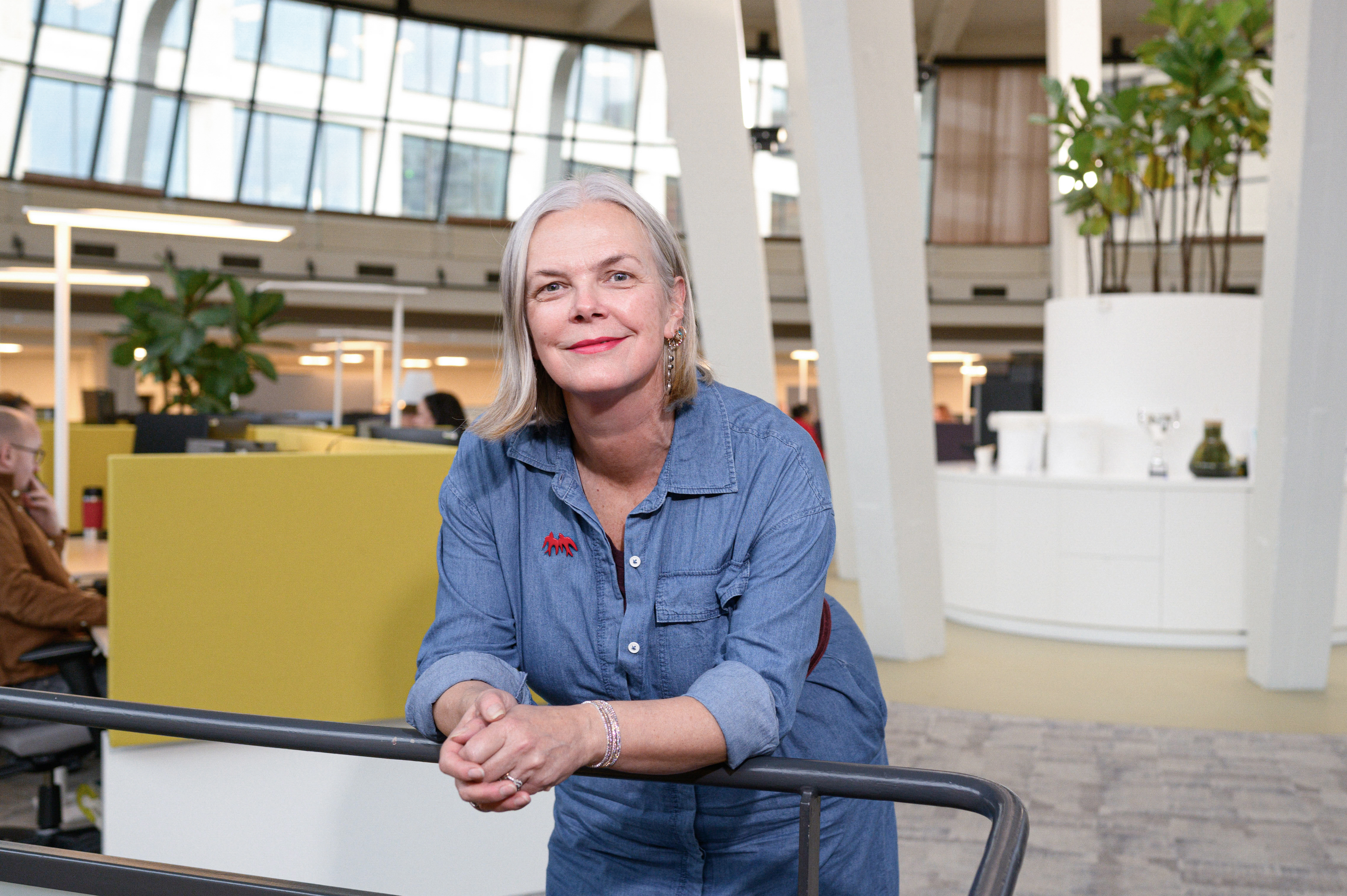 Een vrouw leunt ontspannen op een reling en kijkt recht in de camera, met een rustige en zelfverzekerde uitstraling. De achtergrond geeft inzicht in de kantooromgeving.