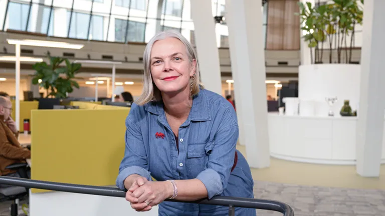 Een vrouw leunt ontspannen op een reling en kijkt recht in de camera, met een rustige en zelfverzekerde uitstraling. De achtergrond geeft inzicht in de kantooromgeving.