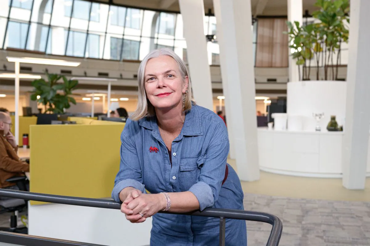 Een vrouw leunt ontspannen op een reling en kijkt recht in de camera, met een rustige en zelfverzekerde uitstraling. De achtergrond geeft inzicht in de kantooromgeving.
