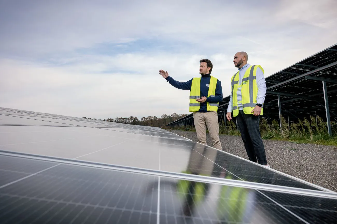 Twee collega’s staan buiten op een veld vol zonnepanelen. Ze zijn met elkaar in gesprek en één van hen wijst naar iets in de verte.